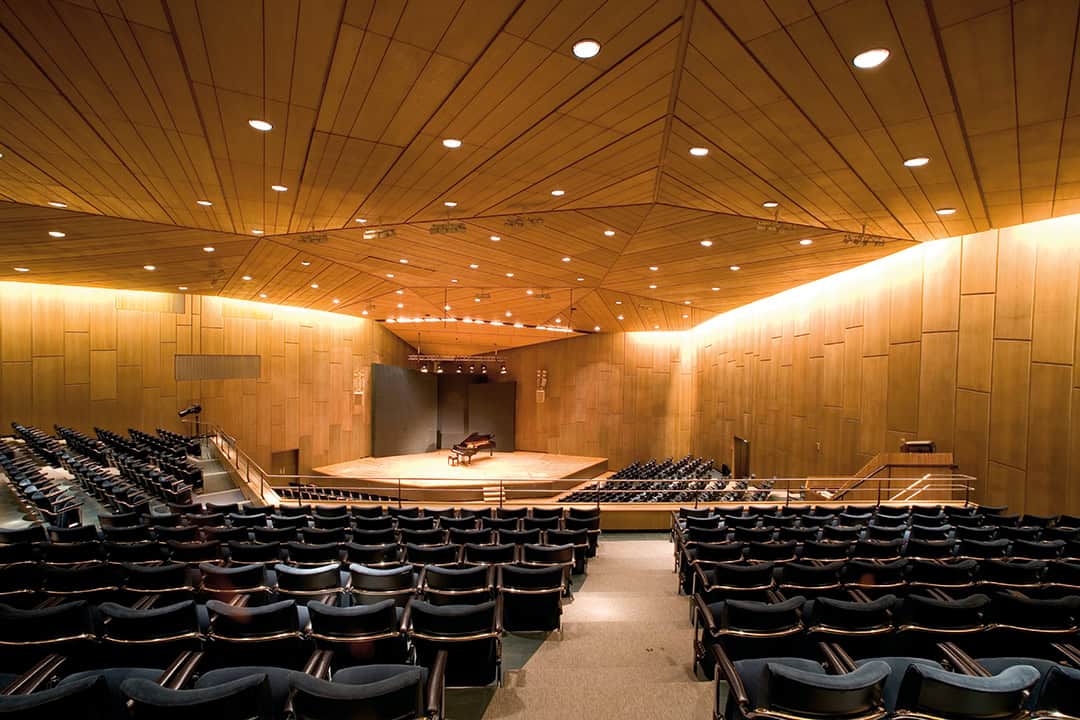 Ein geräumiger Konzertsaal mit geometrischer Holzdecke, leeren türkisfarbenen Sitzen und einem Flügel auf der Bühne. Warmes Licht erhellt den Raum und unterstreicht die moderne Architektur und das Design.