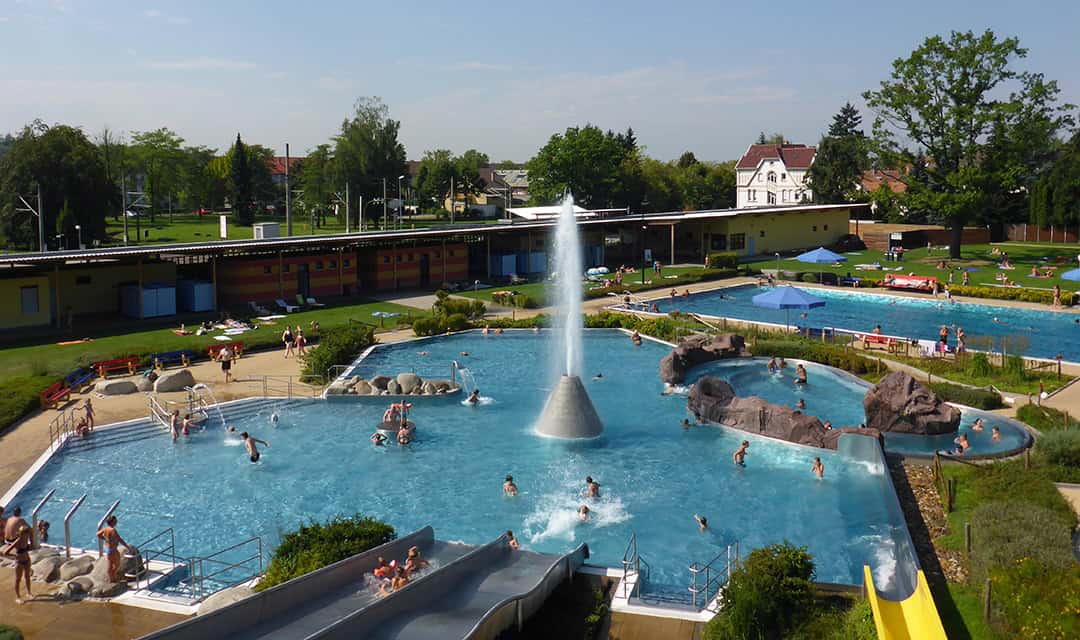 Ein großes öffentliches Freibad mit einem zentralen Springbrunnen, umgeben von schwimmenden und entspannenden Menschen. Der Bereich umfasst Rutschen, Liegeflächen und Grünflächen. Im Hintergrund sind Gebäude und Bäume unter einem klaren Himmel zu sehen.