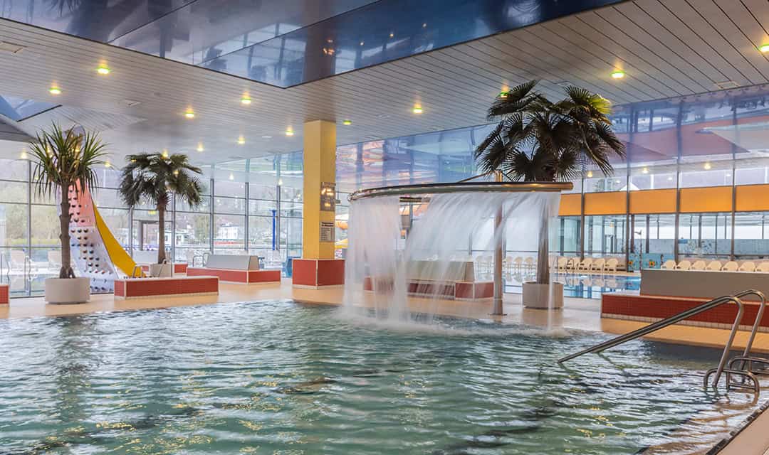 Innenpoolbereich mit Wasserfall und gelber Rutsche. Der Raum ist hell, mit großen Fenstern und künstlichen Palmen. Liegestühle säumen den Hintergrund, und die Decke ist mit reflektierenden Paneelen und Lichtern gestaltet.