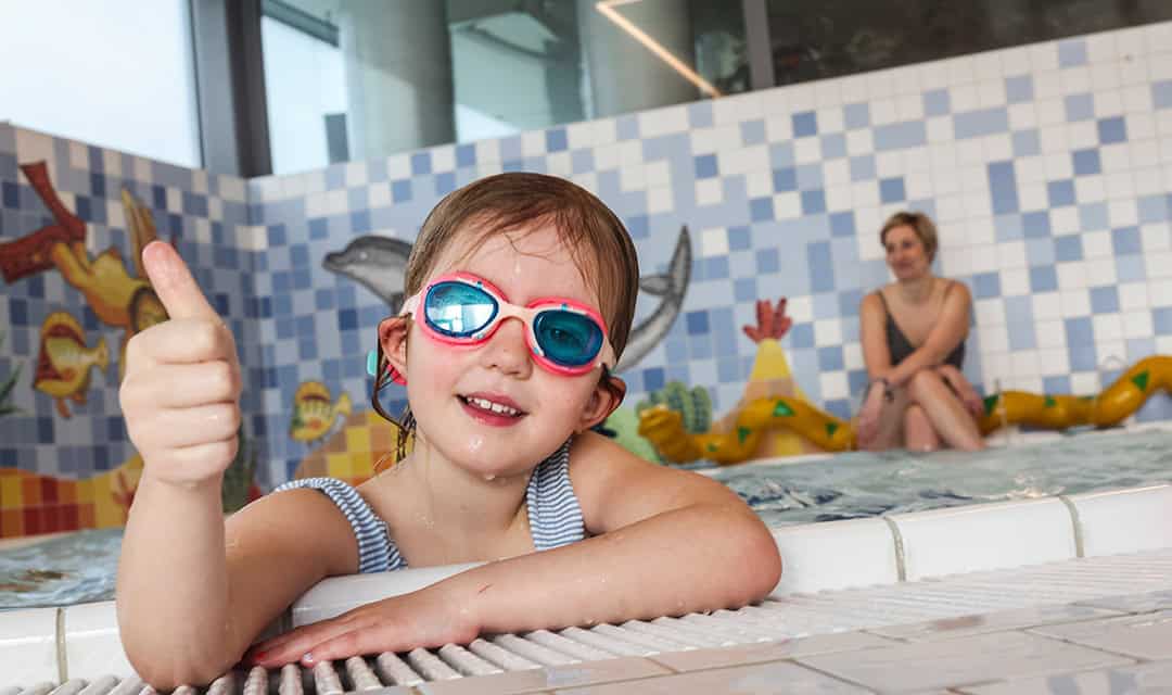 Ein kleines Kind mit Schwimmbrille und gestreiftem Badeanzug lehnt am Beckenrand und streckt den Daumen hoch. Im Hintergrund sitzt eine Frau neben bunten Wandfliesen mit Wassermotiven.