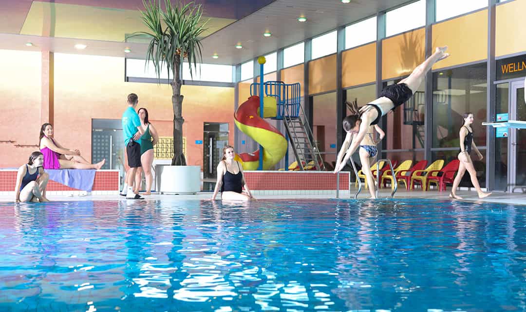 Menschen genießen ein Hallenbad; eine Person springt hinein, andere sitzen am Beckenrand. Im Hintergrund befinden sich eine bunte Spiralrutsche und Liegestühle am Beckenrand. Helle Fenster und eine Topfpflanze verstärken die lebendige Atmosphäre.