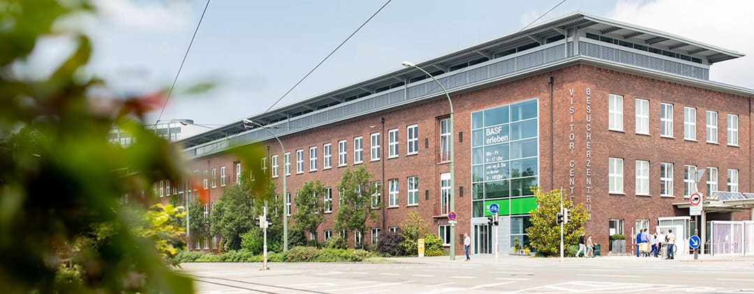 BASF_Besucherzentrum_Aussenansicht_1080 Ein modernes Backsteingebäude mit großen Fenstern und einem Schild mit der Aufschrift „BESUCHERZENTRUM“. Im Vordergrund sind Bäume und Büsche zu sehen, und Menschen gehen in der Nähe des Eingangs spazieren. Der Himmel ist klar, nur vereinzelt bewölkt.