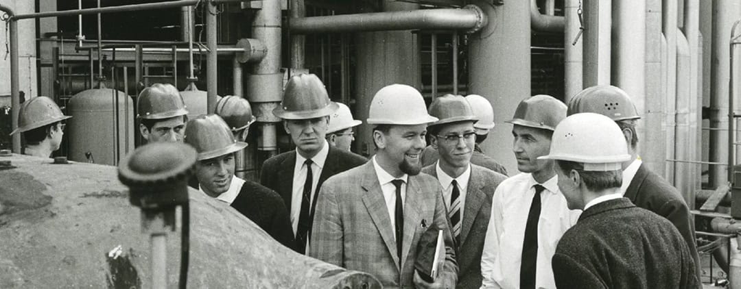 Betriebsbesichtigung_1966_1080 Eine Gruppe von Männern mit Schutzhelmen und Anzügen hat sich auf einem Industriegelände versammelt und unterhält sich. Im Hintergrund sind Industrieanlagen und Rohrleitungen zu sehen, was auf eine Szenerie aus der Mitte des 20. Jahrhunderts hindeutet.