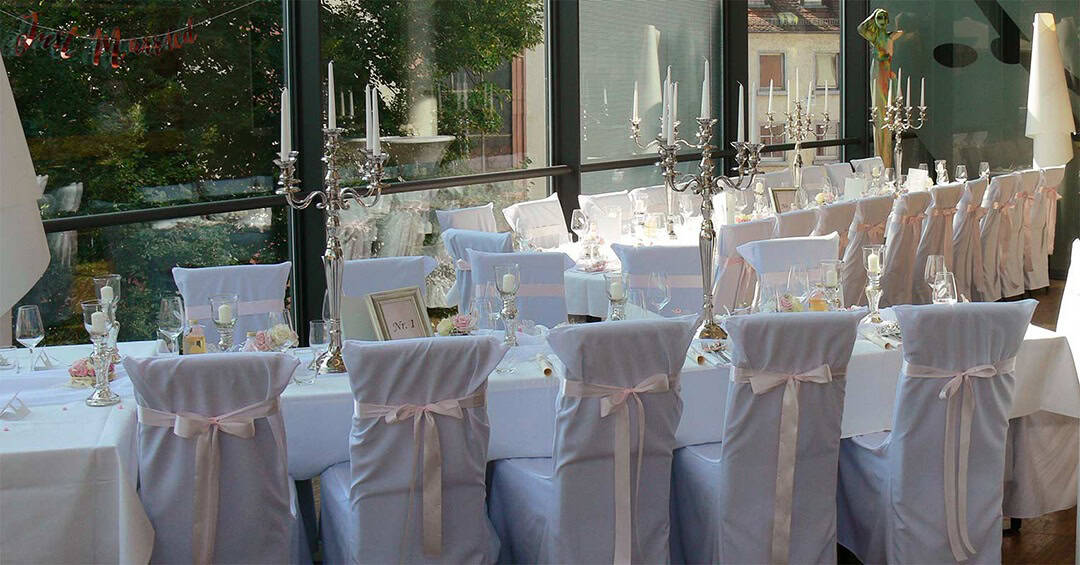 Hochzeit-im-Jedermann-1080 Eleganter Hochzeitsempfang im Innenbereich mit langen, weiß gedeckten Tischen. Die Stühle sind mit weißen Hussen und rosa Bändern bedeckt. Die Tische sind mit hohen Kandelabern und Glaswaren geschmückt und stehen an großen Fenstern mit Blick ins Grüne.