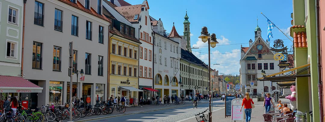 MarcushausAsamRathaus_1080 Eine belebte europäische Straße, gesäumt von historischen Gebäuden, Geschäften und Cafés. Menschen gehen und radeln auf dem Kopfsteinpflaster. Unter blauem Himmel ist ein Uhrturm mit einer bayerischen Flagge zu sehen.