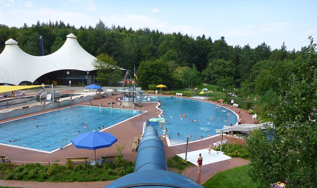 Ein großes Freibad mit Wasserrutsche, umgeben von grünen Bäumen. Schwimmer genießen das Wasser an einem sonnigen Tag unter blauen Sonnenschirmen und verstreuten Liegestühlen. Im Hintergrund sind Zelte und ein Waldgebiet zu sehen.