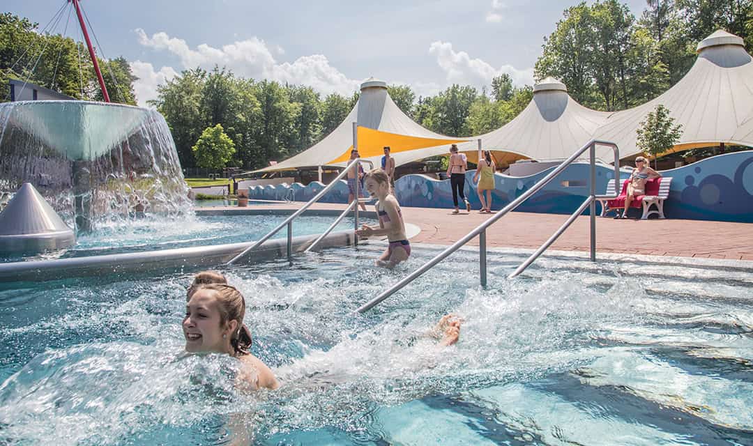 Kinder spielen und schwimmen in einem Außenpool mit Wasserspielen. Ein Stoffdach und Bäume im Hintergrund spenden Schatten. Der Himmel ist klar und sonnig und sorgt für eine lebendige und fröhliche Atmosphäre.