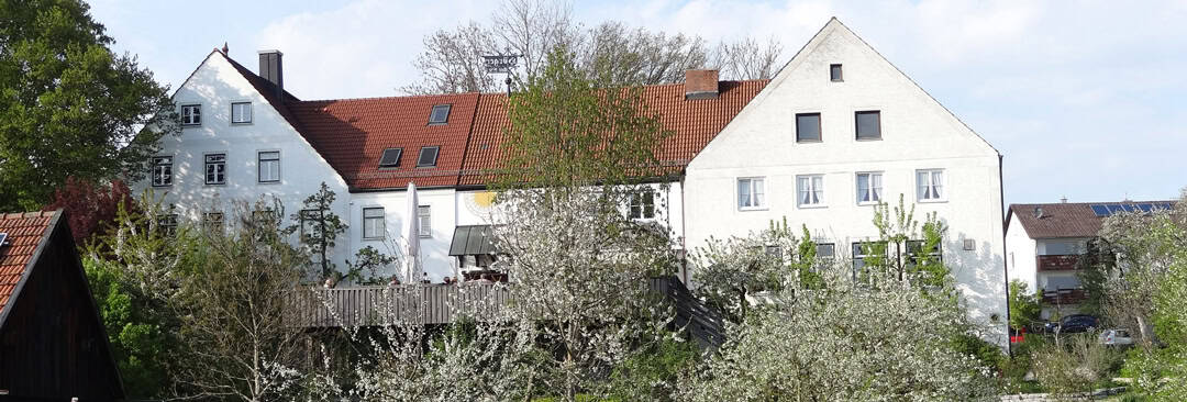 aussenansicht_suedseite_gast_tafernwirtschaft_hoerger Ein großes weißes Haus mit rotem Ziegeldach wird teilweise von blühenden Bäumen und Sträuchern verdeckt. Der Himmel ist klar und im Hintergrund ist ein kleineres Gebäude zu sehen.