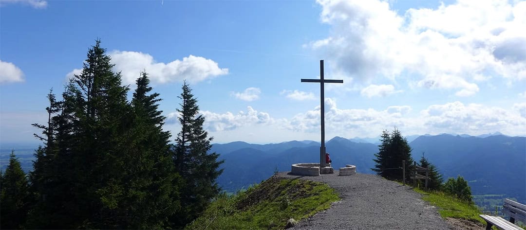brauneck_1080 Ein großes Kreuz steht auf einem Schotterweg auf einem Berggipfel und bietet Ausblick auf eine Reihe blauer Berge unter einem Himmel mit vereinzelten Wolken. Hohe immergrüne Bäume säumen den Weg, und eine Person sitzt in der Nähe des Kreuzes und genießt die Aussicht.