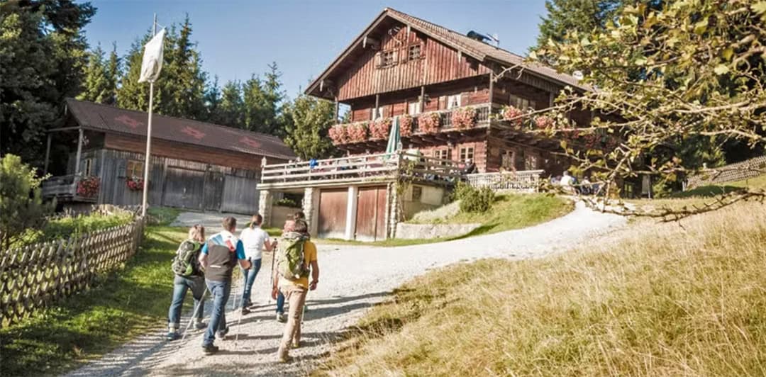 denkalm_1080 Eine Gruppe von Menschen geht auf ein rustikales Holzhaus zu, das mit Blumenkästen geschmückt ist. Das Gebäude liegt inmitten einer üppigen, grünen Landschaft mit Bäumen und einem Fahnenmast in der Nähe. Ein Zaun säumt den Weg, und rechts erstreckt sich ein grasbewachsener Hügel.