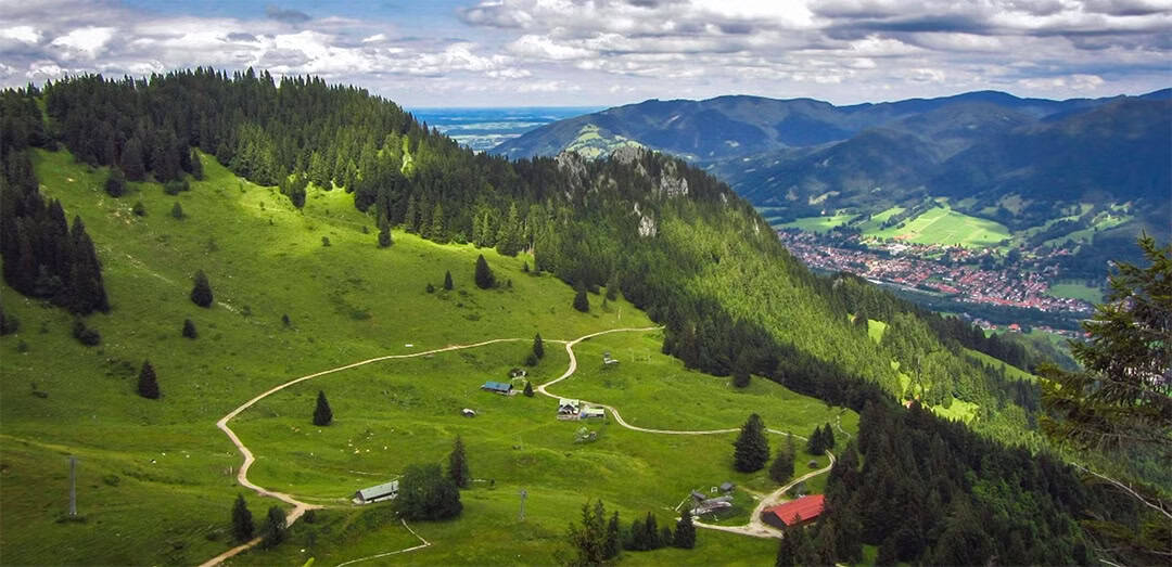 lenggries_wandern_1080 Luftaufnahme eines üppig grünen Tals mit gewundenen Pfaden, kleinen Gebäuden und vereinzelten Bäumen. Auf der einen Seite erheben sich bewaldete Hügel, und in der Ferne liegt eine Stadt zwischen Bergen unter einem teilweise bewölkten Himmel.