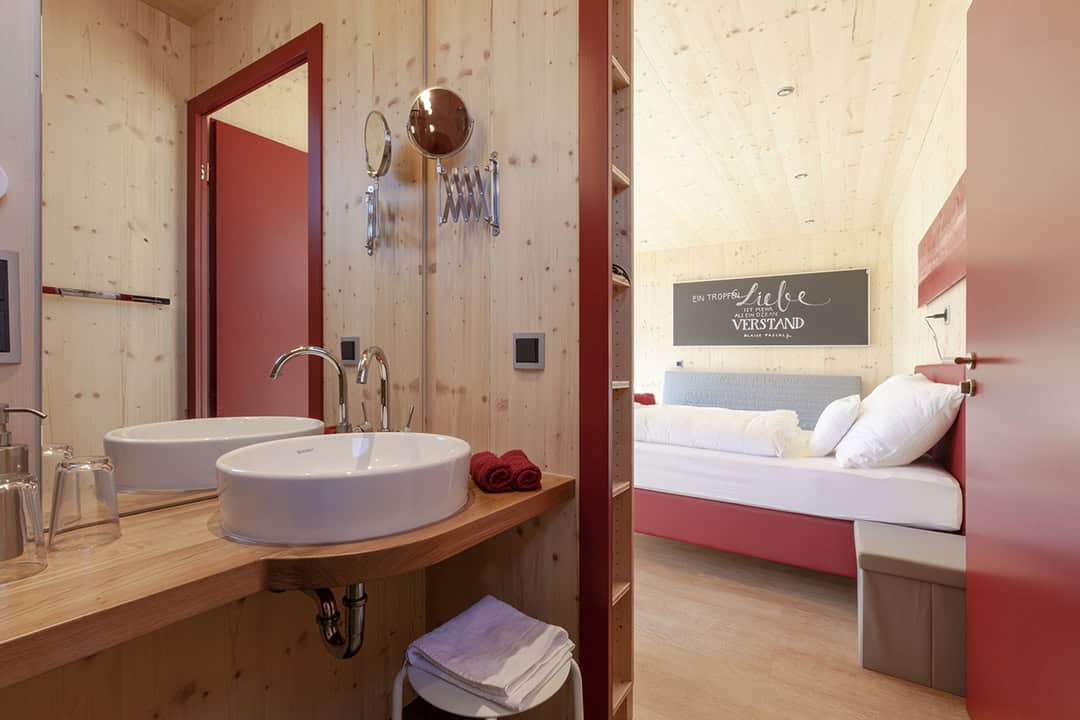 Deluxe-Studio-C---Bad-1080 Ein gemütliches Holzzimmer mit einem Badezimmer im Vordergrund, ausgestattet mit einem Waschbecken und einem runden Spiegel. Im Hintergrund steht ein ordentlich gemachtes Bett mit weißen Kissen. Eine Wanddekoration zeigt einen Text in stilvoller weißer Schrift.