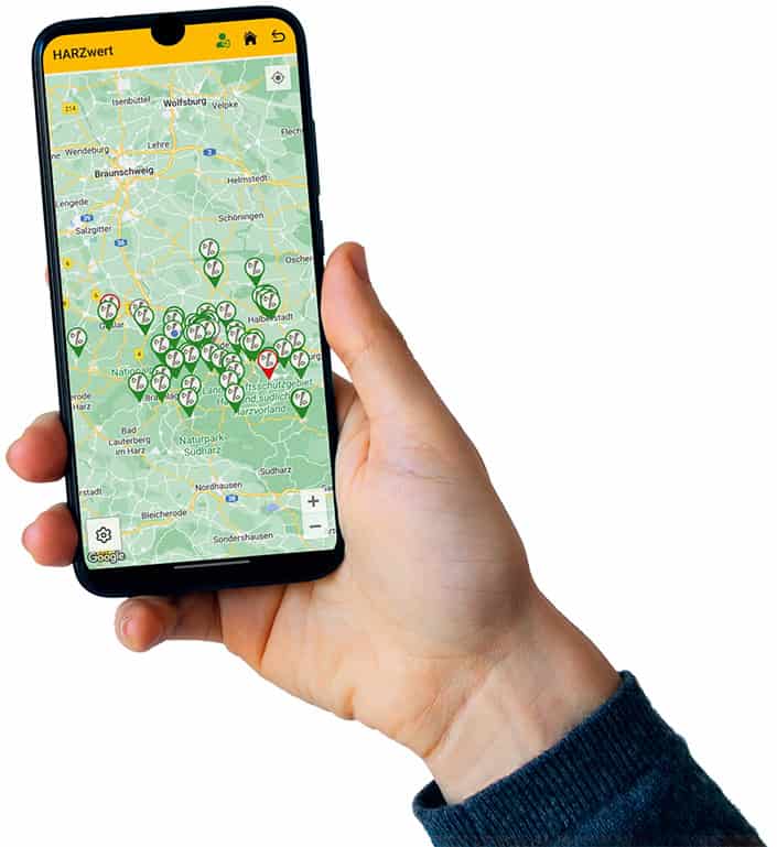 HARZwert_Handy_mit_Karte_705 Eine Hand hält ein Smartphone mit einer Karte und mehreren Standortmarkierungen. Die Karte scheint auf eine Region mit markierten Städten und Autobahnen fokussiert zu sein. Die Telefonoberfläche zeigt oben ein grünes Logo und verschiedene Symbole.