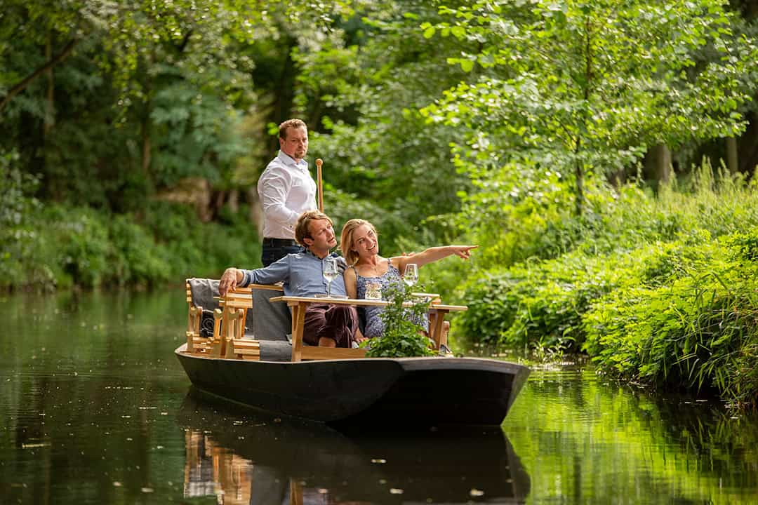 Kahntour-auf-den-Spreewaldfließen_(c)Amt-Burg-(Spreewald),-Ron-Petraß_1080 Ein Paar sitzt mit einem Glas Wein auf einem kleinen Holzboot, während eine Person das Boot durch einen üppigen, grünen Kanal steuert. Die Frau deutet in die Ferne, und die Atmosphäre ist ruhig und entspannt.