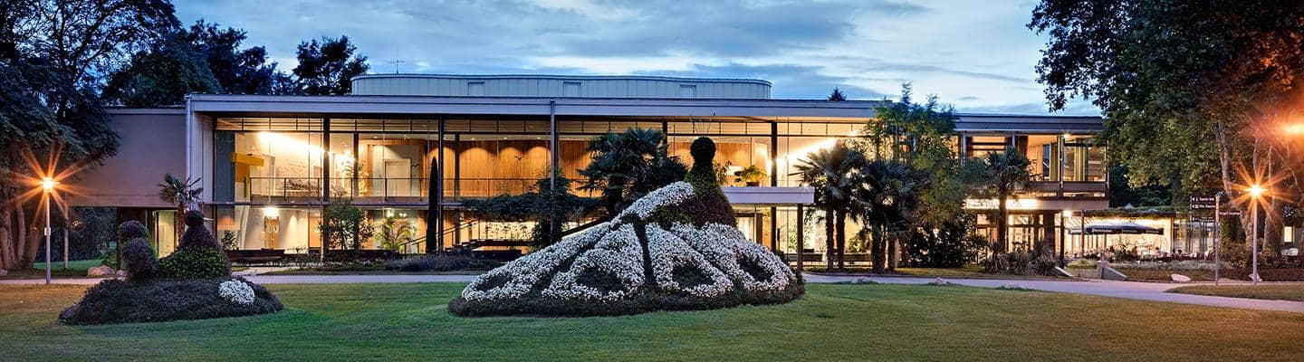 Kurhaus-Bad-Krozingen Ein modernes Gebäude mit großen Fenstern steht vor einem Abendhimmel. Im Vordergrund sind kunstvoll geformte Hecken mit Blüten, die an Pfauen erinnern, umgeben von Gras und Bäumen. Warmes Licht erhellt die Szene.