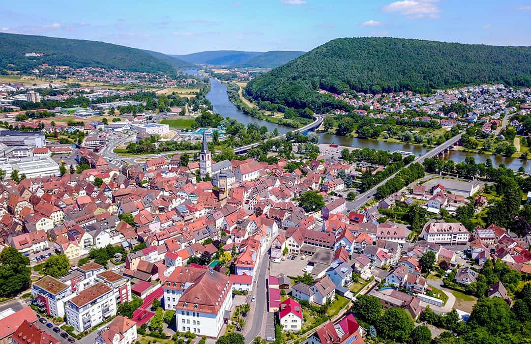 Lohr_Luftaufnahme_Marc-Zeitler_1080 Luftaufnahme einer malerischen Stadt mit rot gedeckten Gebäuden, eingebettet zwischen einem gewundenen Fluss und üppig grünen Hügeln. Eine Brücke überspannt den Fluss und verbindet zwei Stadtteile. Die Landschaft erstreckt sich bis in die fernen Hügel unter einem klaren blauen Himmel.