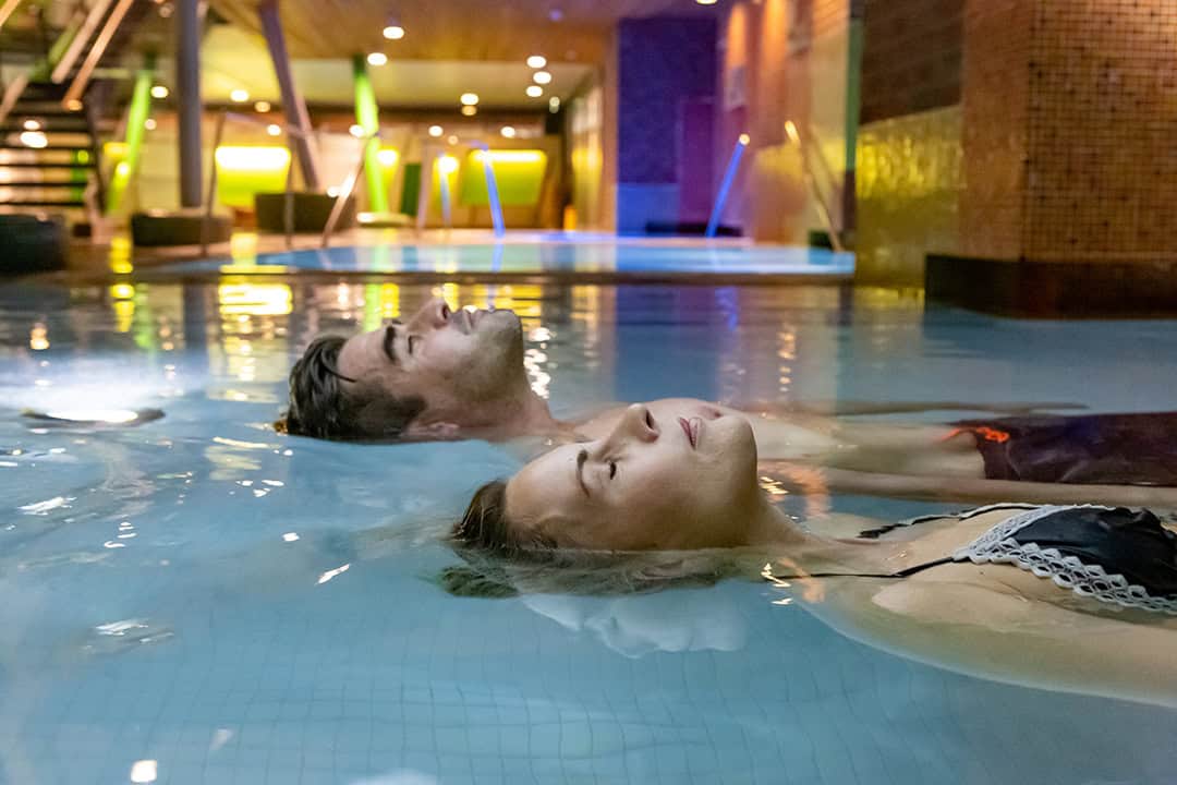 Paar-Sole-Wellness-Schwebebecken-01-_cSpreewald-Therme-GmbH_-Fotografin-Beate-Waetzel_1080 Zwei Menschen entspannen sich in einem schwach beleuchteten Hallenbad und treiben mit geschlossenen Augen auf dem Rücken. Die stimmungsvolle Beleuchtung wirft einen ruhigen Schein auf das Wasser. Im Hintergrund sind Treppen und eine Backsteinmauer zu sehen.