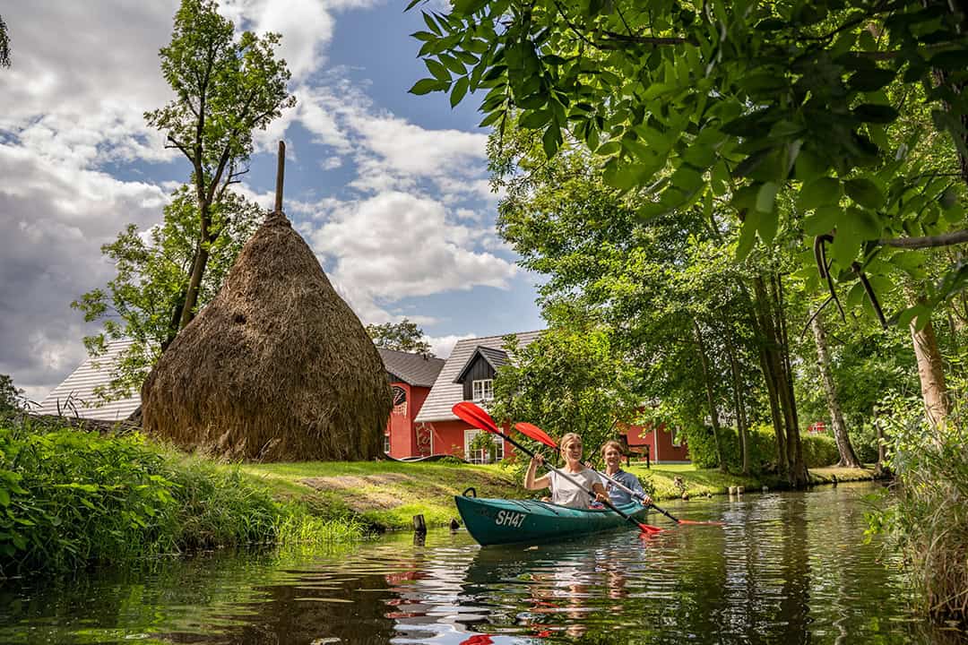 Paddeltour-durch-die-Streusiedlung_(c)Amt-Burg-(Spreewald),-Ron-Petraß_1080 Zwei Personen paddeln in einem Kanu auf einem schmalen, von Bäumen gesäumten Wasserweg. In der Nähe befinden sich ein rustikaler, strohgedeckter Heuhaufen und farbenfrohe Häuser. Die Szenerie ist üppig und grün, darüber ein teilweise bewölkter Himmel.