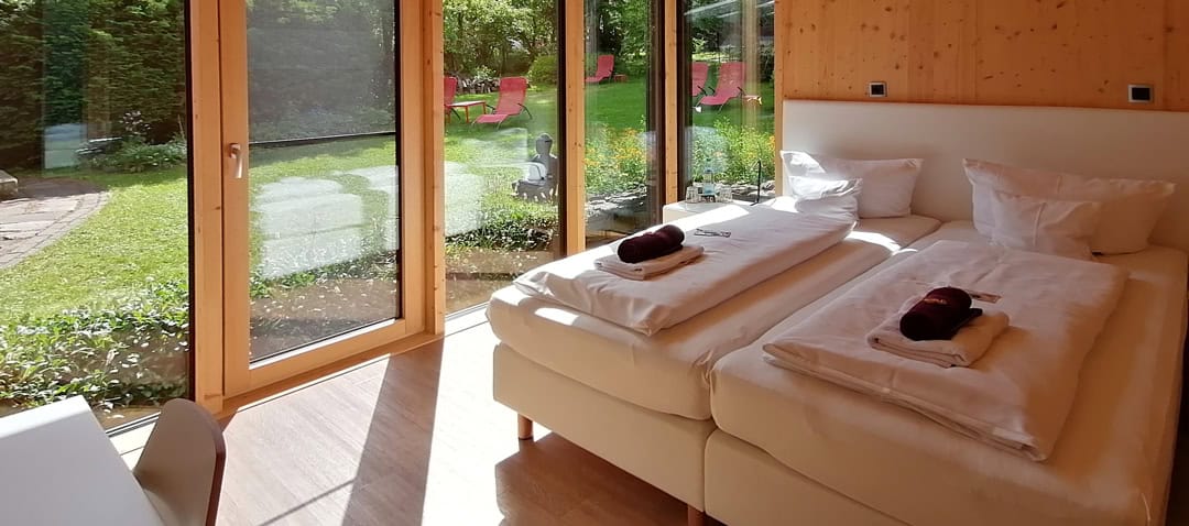 Zimmer_1080 Helles Zimmer mit zwei sauber bezogenen, zusammengeschobenen Einzelbetten, jeweils mit Handtuch und Toilettenartikeln. Große Glastüren öffnen sich zu einem üppigen Garten mit mehreren roten Liegestühlen. Holzböden und helle Holzvertäfelungen sorgen für eine gemütliche Atmosphäre.