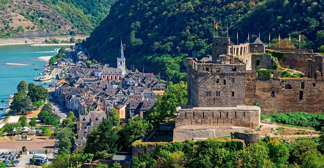 ansicht-st-goar_1080 Malerischer Blick auf eine malerische Stadt am Flussufer mit einer historischen Steinburg auf einem Hügel. Die Stadt besticht durch charmante Gebäude und einen hohen Kirchturm, eingebettet zwischen üppig grünen Hügeln und einem gewundenen Fluss unter einem klaren blauen Himmel.
