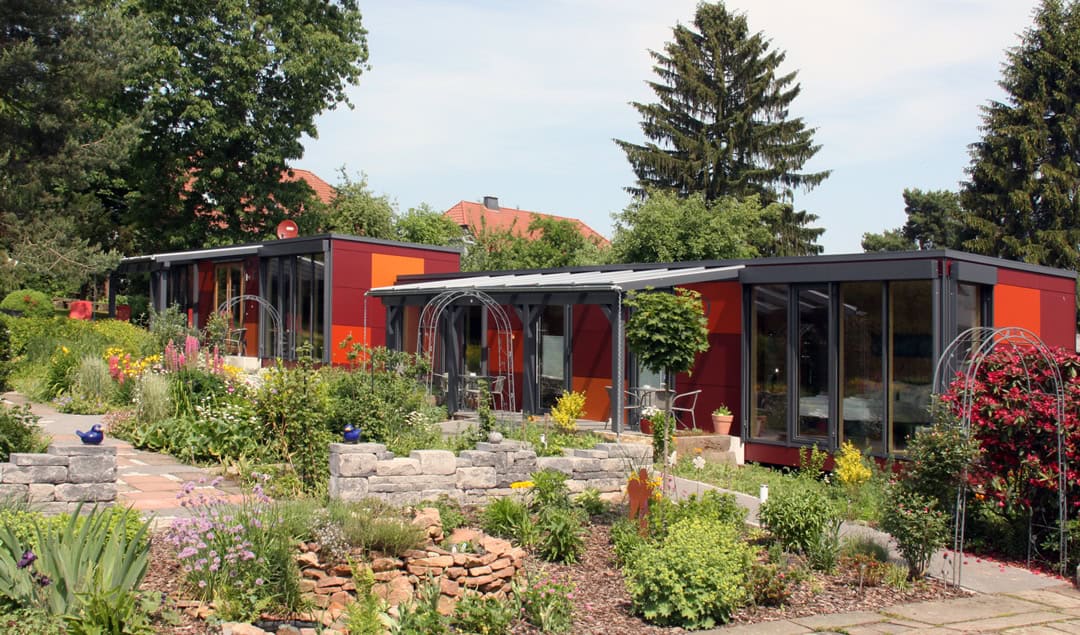kleinhotel_garten-1080 Moderne rote Modulgebäude mit großen Glasfenstern in einem üppigen Garten. Steinwege und Blumenbeete mit leuchtend gelben und violetten Blüten stehen im Vordergrund, im Hintergrund hohe Bäume unter blauem Himmel.