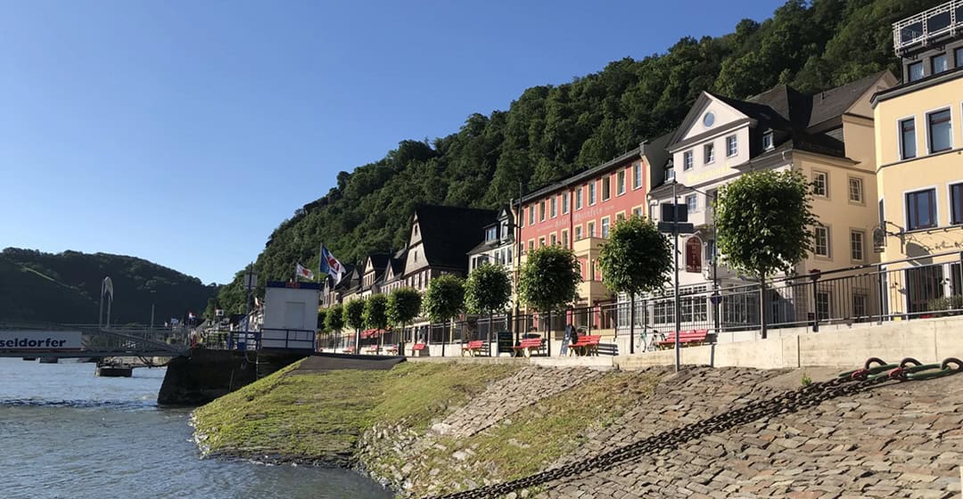 uferpromenade-in-st-goar_1080 Eine malerische Flusslandschaft zeigt eine Reihe farbenfroher Gebäude mit Giebeldächern, gesäumt von Bäumen. Ein ruhiger Fluss fließt vorbei, gesäumt von einem grasbewachsenen Uferdamm. An der Promenade genießen Menschen den sonnigen Tag.