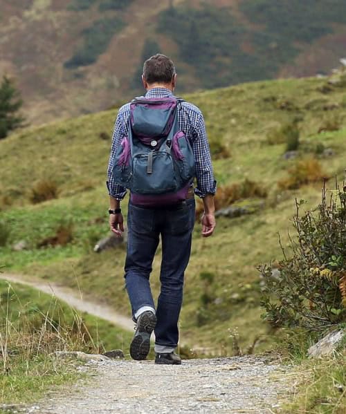 Eine Person in kariertem Hemd, Jeans und Rucksack geht einen schmalen Pfad durch eine grasbewachsene, hügelige Landschaft entlang. Das Gelände ist mit Sträuchern und Felsen übersät, im Hintergrund sind ein Baum und Hügel zu sehen.