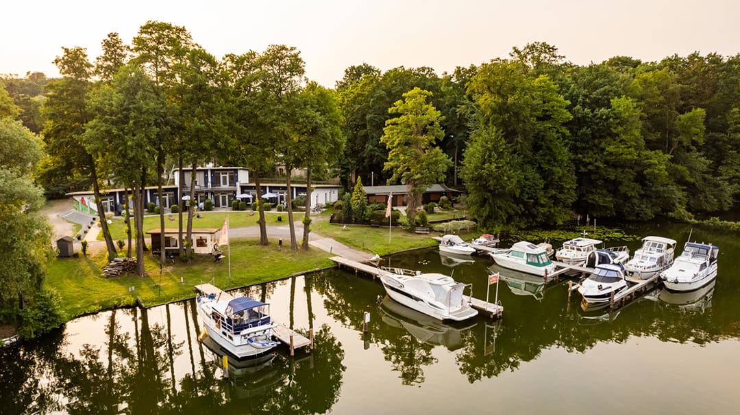 Ferienpark_Porta_Elisa_Artem_Heissig-2_1080 Eine ruhige Seeszene mit mehreren Booten, die in einem von üppigen Bäumen umgebenen Yachthafen anlegen. Am Ufer ist ein Gebäude mit Terrasse zu sehen, und das Wasser spiegelt das Grün und die Boote wider, was eine friedliche Atmosphäre schafft.
