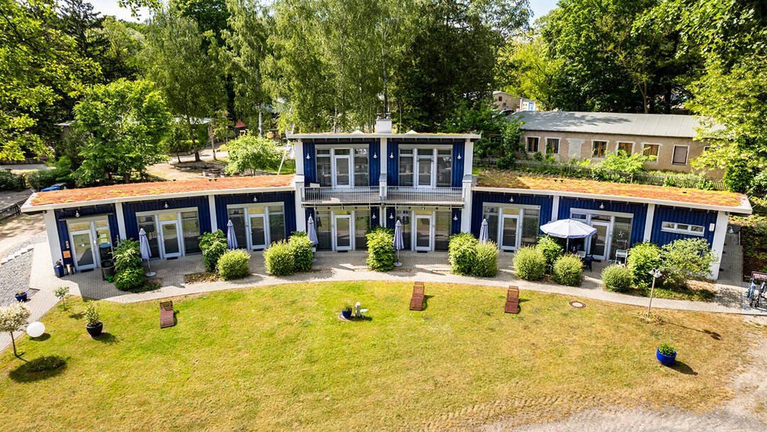 Ferienpark_Porta_Elisa_Artem_Heissig-3_1080 Ein modernes, einstöckiges, blaues Gebäude mit einem zentralen Obergeschoss, umgeben von Grünflächen. Das Dach ist mit Rasenflächen bepflanzt und verfügt über Gartenstühle und Pflanzen. Das Gebäude verfügt über große Fenster und liegt in einer ruhigen, waldreichen Umgebung.
