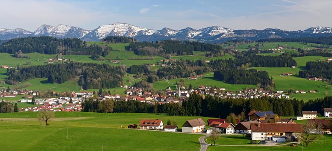 Panorama_Weiler-Simmerberg_1080 Ein üppig grünes Tal mit verstreuten Häusern und kleinen Städten, umgeben von sanften Hügeln. Schneebedeckte Berge unter einem klaren blauen Himmel bilden den Hintergrund, während Bäume und Felder den Vordergrund füllen.