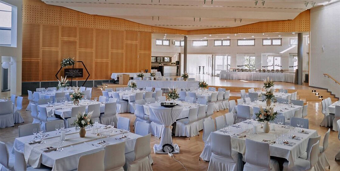 BaltKult_Hochzeit_1080 Ein geräumiger Bankettsaal mit mehreren runden, weiß gedeckten Tischen, elegant arrangiert mit floralen Tischdekorationen. Der Holzboden bildet einen Kontrast zum modernen Interieur, und große Fenster auf der rechten Seite lassen viel Tageslicht herein.