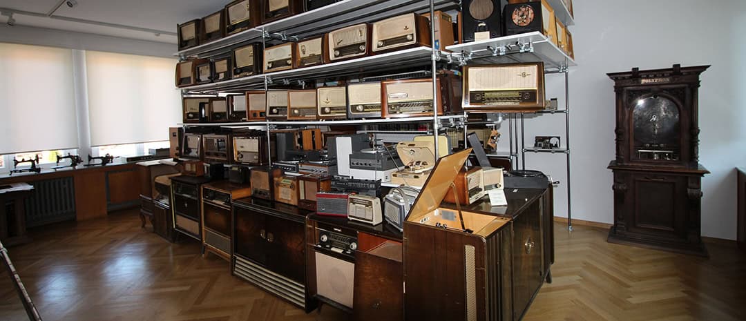Ein Raum voller Vintage-Radios und Plattenspieler, ausgestellt auf Regalen und Holzschränken. Die Sammlung variiert in Größe und Design und verfügt über eine Vielzahl von Knöpfen und Zifferblättern. In der Ecke steht eine große, kunstvolle Uhr auf einem Holzboden.