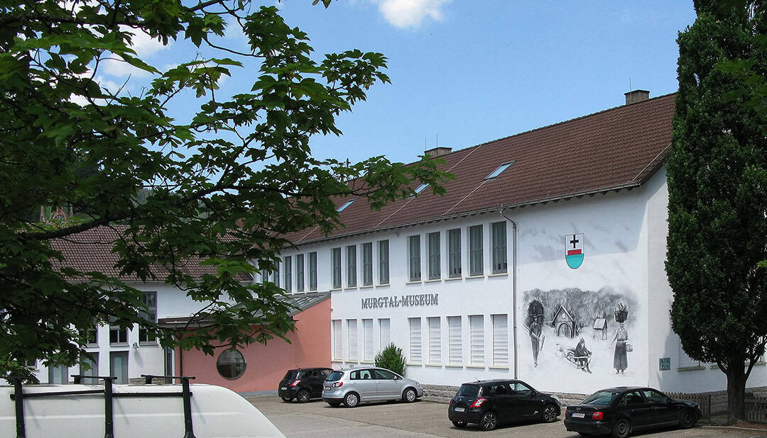 Ein zweistöckiges Gebäude mit weißen Wänden und braunem Dach, das mit einem Wandgemälde und einem Wappen mit einem roten Kreuz geschmückt ist. Mehrere Autos parken davor, und links rahmt ein Baum die Szene teilweise ein. Der Himmel ist blau mit vereinzelten Wolken.