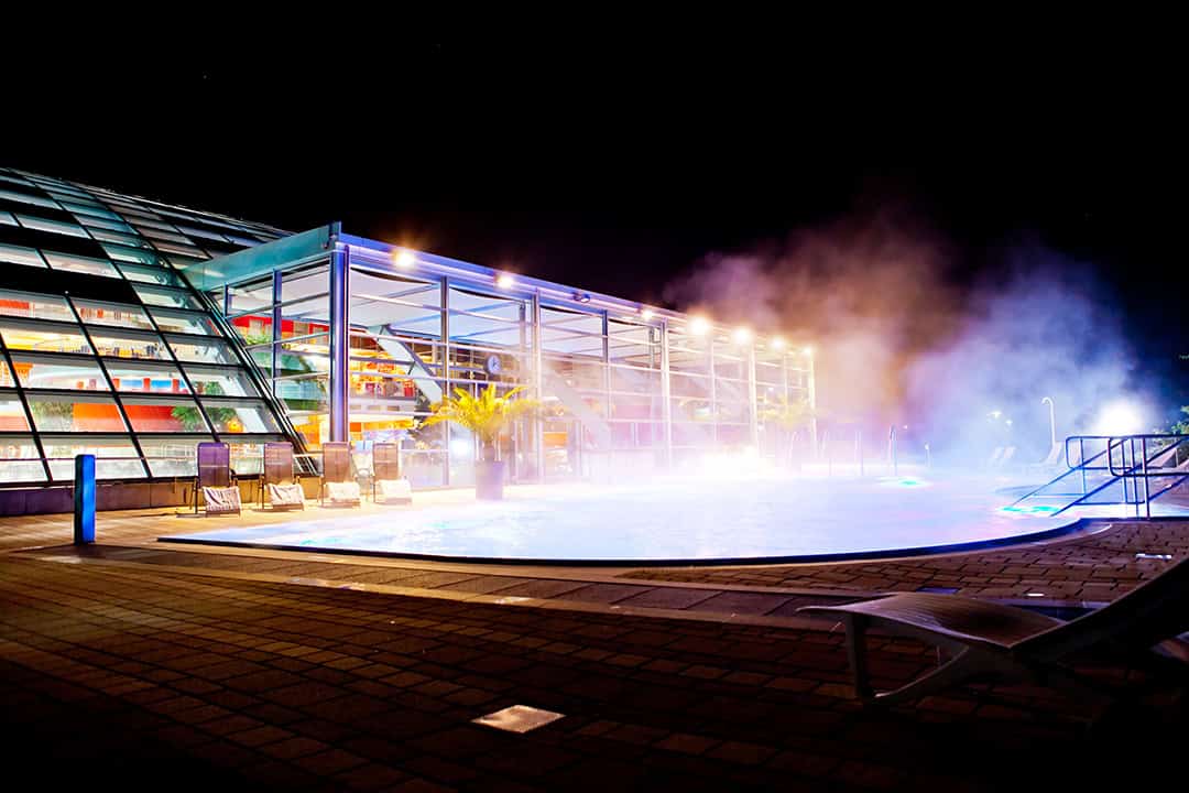 cm_Badewelt-Nacht_1080 Ein hell erleuchteter, beheizter Außenpool, umgeben von einer modernen Glaskonstruktion, dampft aus dem Wasser und rundherum stehen Liegestühle. Das Ambiente ist warm und einladend.