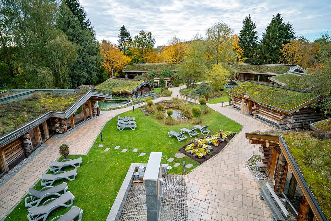 cm_saunagarten_1080 Ein ruhiger Spa-Bereich im Freien mit Gebäuden im Blockhausstil und grünen Dächern. Gepflasterte Wege, Liegestühle, gepflegte Gärten und herbstliche Bäume unter leicht bewölktem Himmel runden das Ambiente ab.