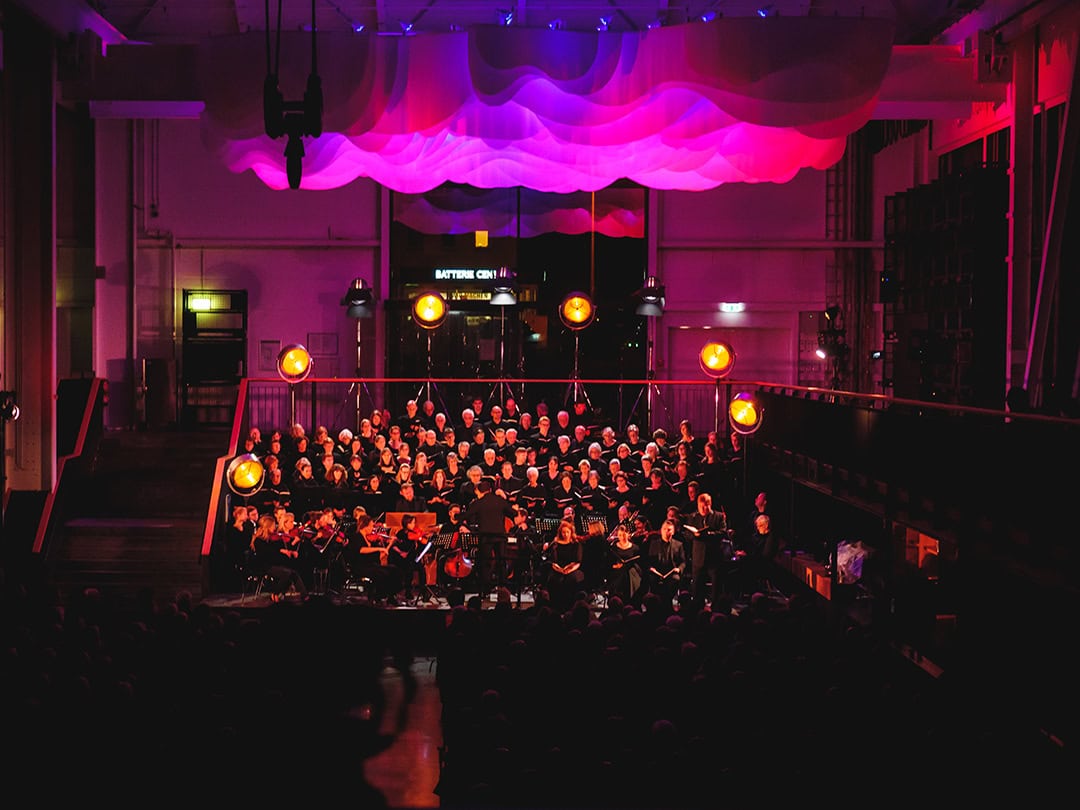 mood-of-Mozart_1080 Ein Chor und ein Orchester treten auf einer schwach beleuchteten Bühne vor einem leuchtend violetten und roten Hintergrund auf. Kreisförmige Lichter leuchten die Musiker an. Die Szene spielt in einem großen Saal und schafft eine bezaubernde Atmosphäre.