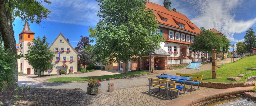 Dobel__Kirche__Schulhaus__Rathaus__1080 Charmante Dorfszene mit farbenfrohen Gebäuden, einem zentralen Weg, üppigen Bäumen und einem kleinen Brunnen. In der Nähe steht eine gelb-blaue Bank. Rot gedeckte Gebäude, darunter ein Kirchturm, unterstreichen die malerische Kulisse unter einem leicht bewölkten Himmel.