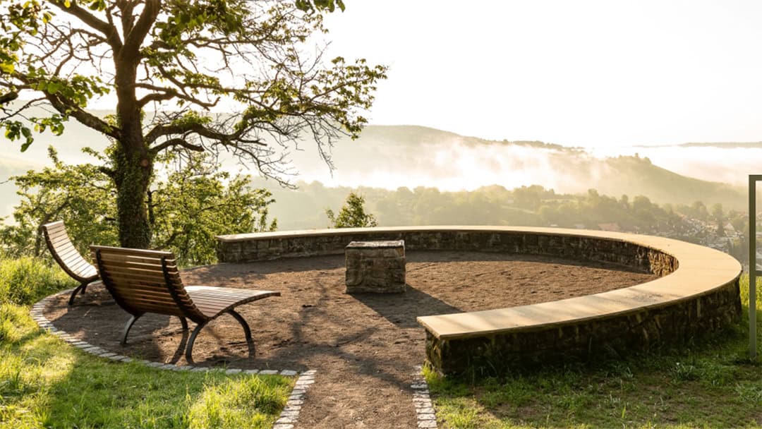 Remshalden_Aussicht_1080 Zwei geschwungene Holzbänke auf einer runden Steinplattform blicken auf ein malerisches, nebliges Tal. Links steht ein Baum mit üppigen grünen Blättern. Im Hintergrund sind sanfte Hügel und ein Wechselspiel aus Sonnenschein und Nebel zu sehen.