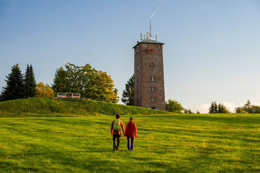 image009_1080 Zwei Menschen gehen Hand in Hand über eine grüne Wiese auf einen hohen, von Bäumen umgebenen Steinturm zu. Der Himmel ist klar, ein Jetstream ist sichtbar. Links neben dem Turm stehen Bänke.
