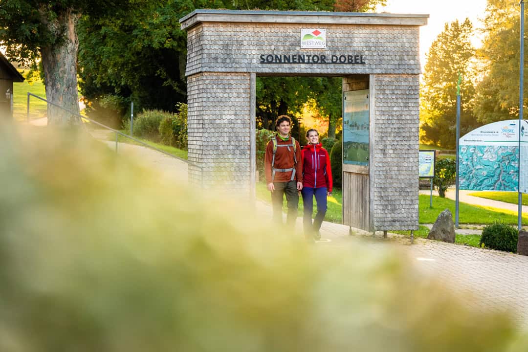 image010_1080 Zwei Personen gehen unter einem hölzernen Torbogen mit der Aufschrift „Sonnentor Dobel“ in einer malerischen, sonnenbeschienenen Gegend hindurch. Sie tragen Outdoor-Kleidung. Im Hintergrund sind Bäume und Schilder zu sehen, die eine Natur- oder Wanderkulisse suggerieren.