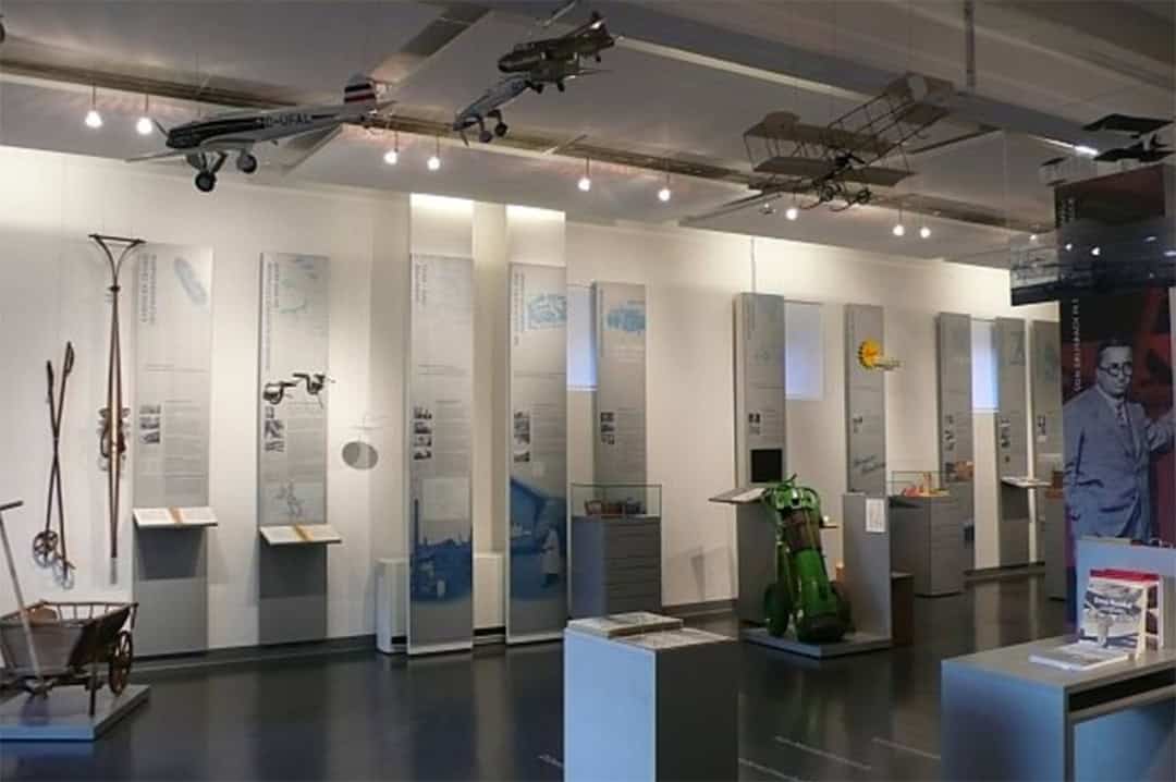 museum_1080 Eine Museumsausstellung zur Geschichte der Luftfahrt mit von der Decke hängenden Modellflugzeugen, Informationstafeln und verschiedenen Artefakten. Der Raum verfügt über einen polierten Boden und eine helle Deckenbeleuchtung.