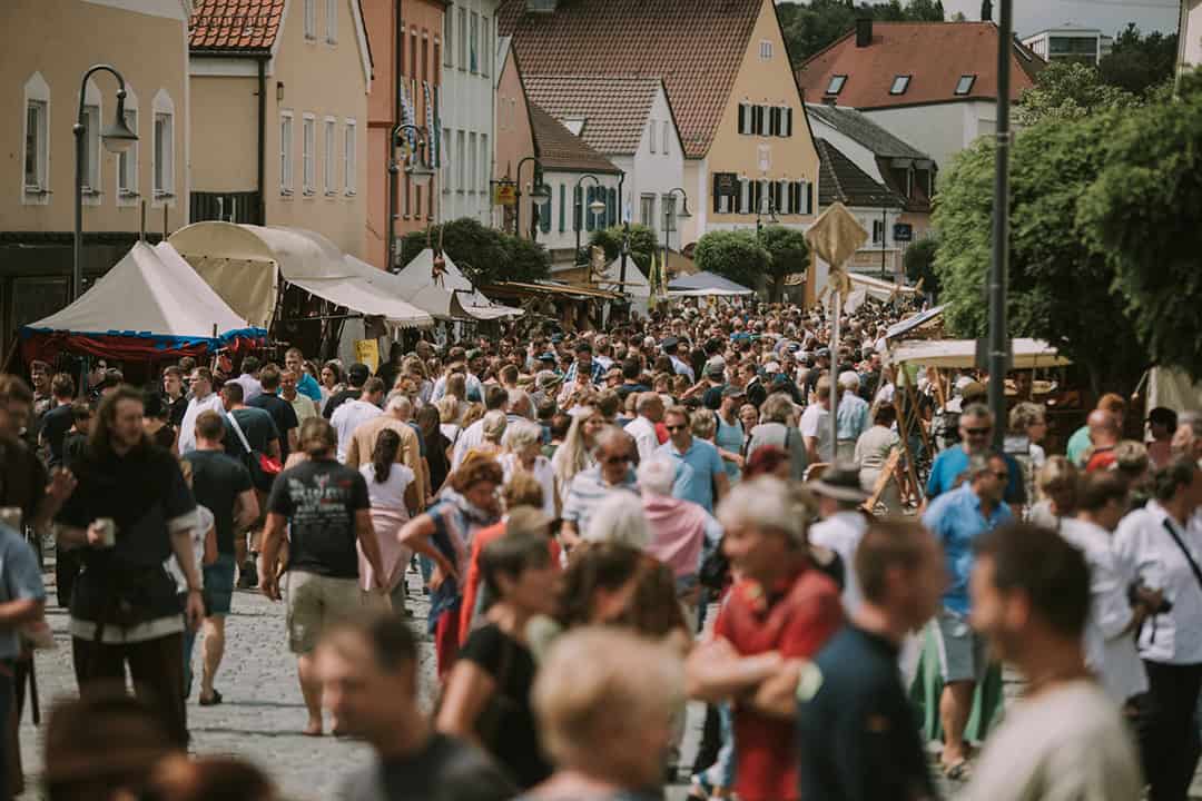 MAF_Arnstorf-(1)_1080 Ein geschäftiges Straßenfest mit einer dichten Menschenmenge, die an Ständen und Geschäften vorbeizieht. Die Straße ist gesäumt von farbenfrohen Gebäuden und Festzelten. Die Atmosphäre ist lebhaft und festlich.