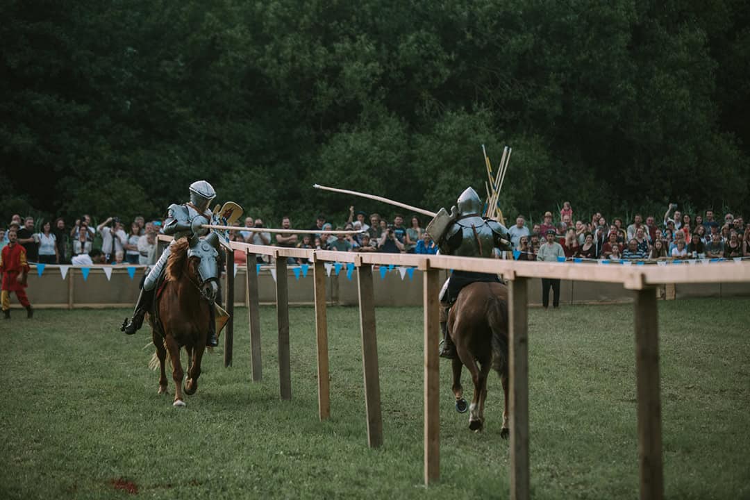MAF_Arnstorf-(2)_1080 Zwei Ritter in Rüstung zu Pferd liefern sich ein Turnier mit Lanzen in einer Freiluftarena. Eine Menschenmenge beobachtet das Geschehen am Rande, vor der Kulisse von Bäumen. Die Szene fängt die Action und Spannung einer mittelalterlichen Nachinszenierung ein.