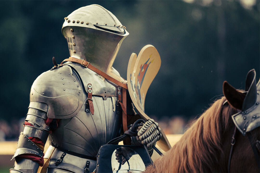 MAF_Arnstorf-(5)_1080 Ein Ritter in glänzender Metallrüstung und Helm reitet auf einem braunen Pferd und hält einen Schild mit bunten Markierungen. Der Hintergrund ist unscharf, was darauf schließen lässt, dass die Szene im Freien stattfindet, möglicherweise bei einer Nachstellung oder einer mittelalterlichen Veranstaltung.