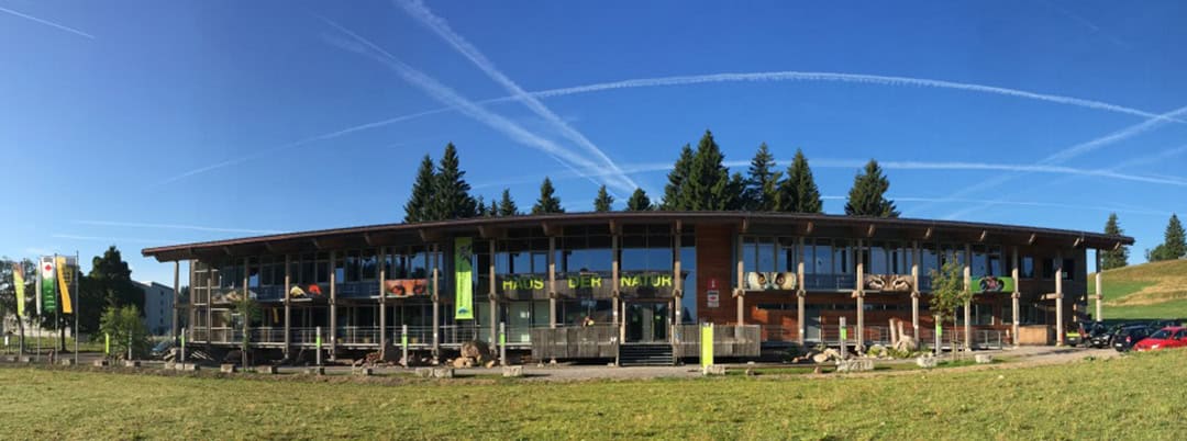 NAZ-Außen-Panorama-1080 Ein modernes Gebäude mit Glasfassade und Holzelementen steht umgeben von Bäumen und Wiesen unter strahlend blauem Himmel. Draußen hängen mehrere Banner, und darüber sind Kondensstreifen von Flugzeugen zu sehen. Rechts vom Gebäude parken Autos.