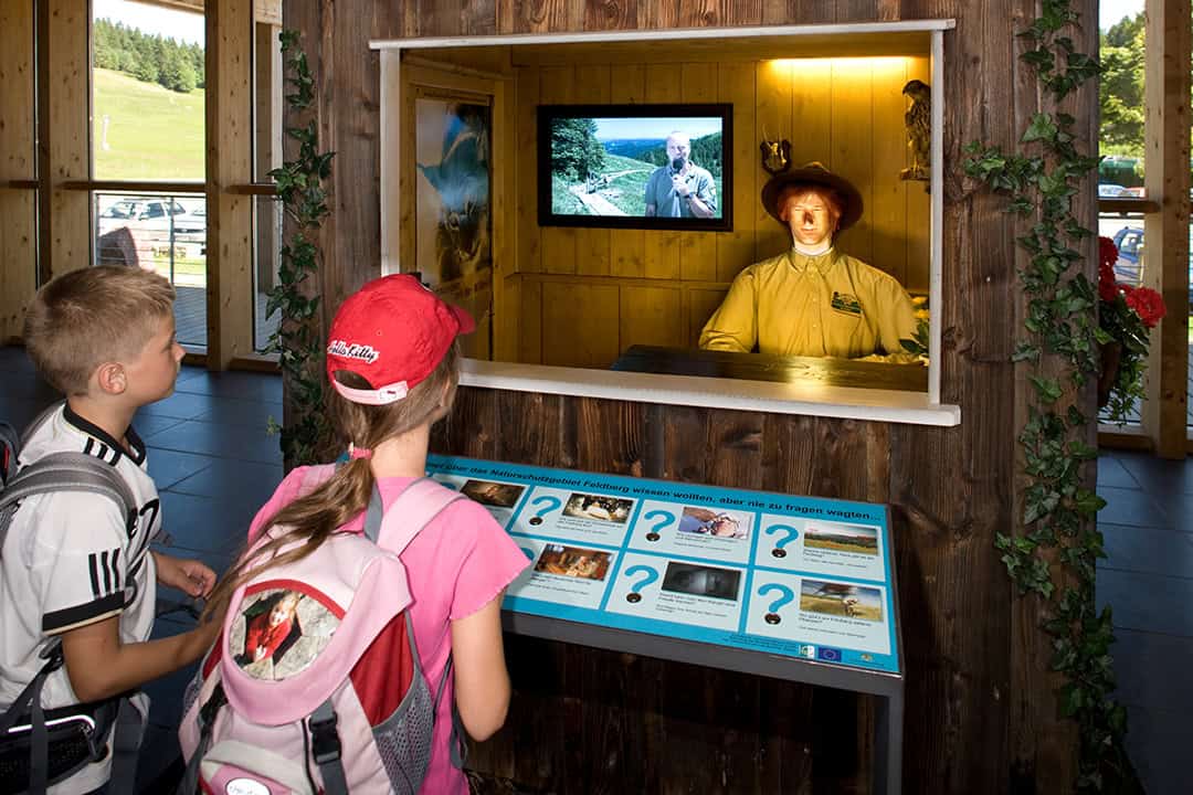 Talking-Ranger-Gocke-1080 Zwei Kinder mit Rucksäcken stehen vor einer interaktiven Ausstellung, die eine animatronische Figur im Ranger-Outfit zeigt. Darüber läuft ein Bildschirm, der ein Video einer Person in der Natur zeigt. Die Szene spielt im Innenbereich, und Informationstafeln sind sichtbar.