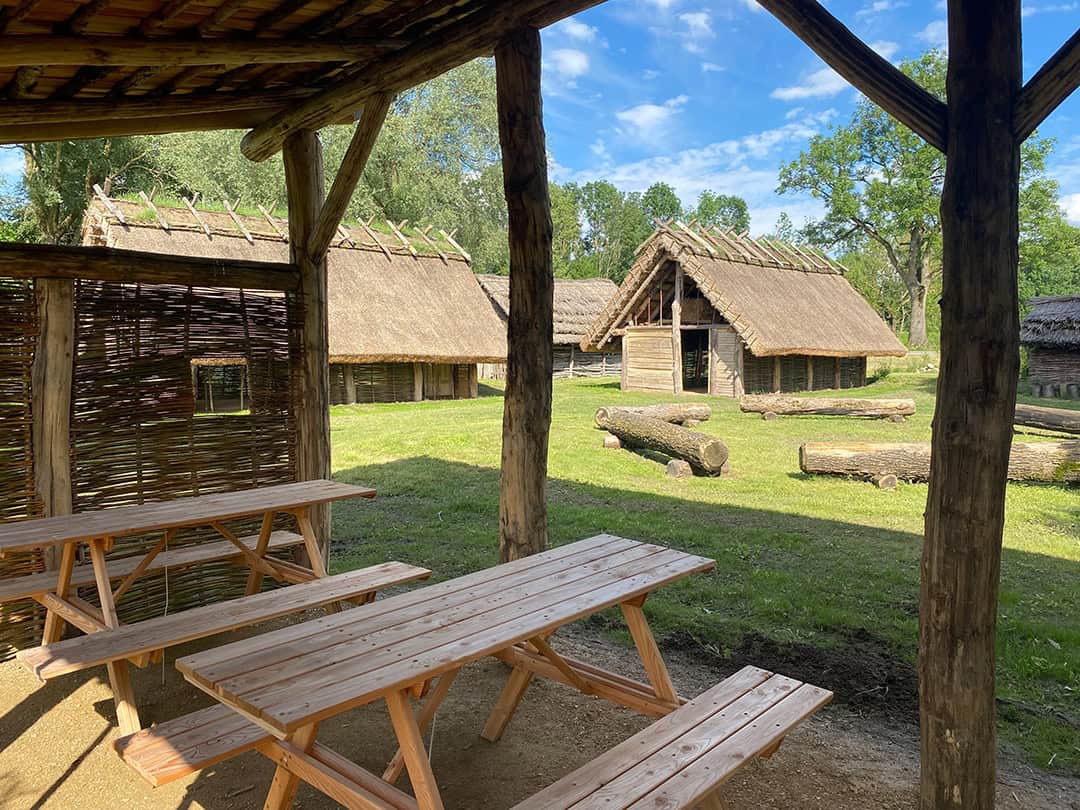 Eine rustikale Szene mit hölzernen Picknicktischen unter einem Baumstammdach mit Blick auf eine Rasenfläche mit mehreren Strohdachhütten. Baumstämme liegen verstreut auf dem Boden, Bäume und ein blauer Himmel prägen den Hintergrund.
