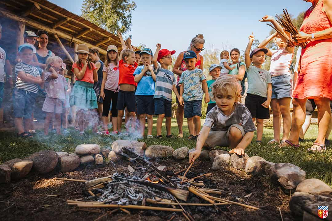 Eine Gruppe von Kindern und Erwachsenen versammelt sich draußen um ein kleines Lagerfeuer. Ein Kind hockt vorn und hält Stöcke über das Feuer. Einige Kinder tragen Hüte und farbenfrohe Kleidung. Alle wirken engagiert und aufmerksam.