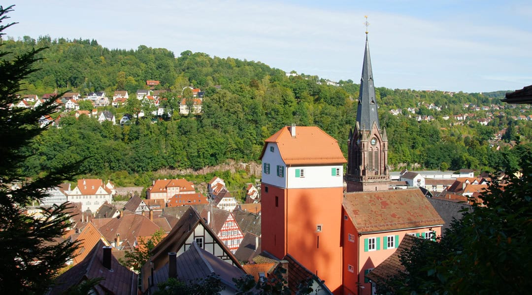 Calw_Der-Lange-und-Stadtkirche-Stadtkirche_Foto-Stadt-Calw_1080 Eine malerische Stadtlandschaft mit rot gedeckten Gebäuden und einer markanten Kirche mit hohem Turm im Vordergrund. Saftig grüne Hügel umgeben die Stadt, und der Himmel ist klar. Die Szenerie ist in helles Sonnenlicht getaucht.