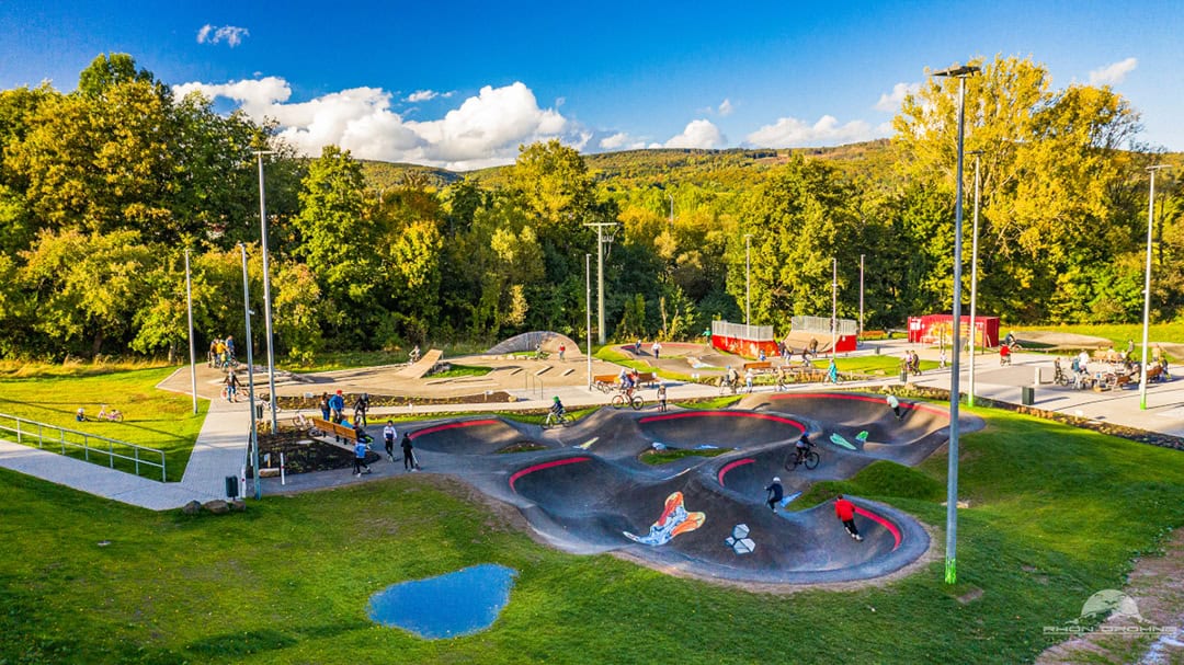 Funpark_1080 Ein lebhafter Skatepark mit Radfahrern und Skatern auf einer kurvigen Strecke inmitten üppiger Vegetation. Hohe Bäume und ein paar Wolken sind im Hintergrund zu sehen. Mehrere Menschen haben sich auf einem gepflasterten Weg versammelt. Rundherum gibt es Bänke und Straßenlaternen.